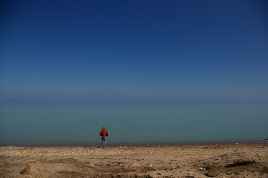 A lone woman in a red coat stands on a sandy beach looking out at the still blue ocean and the dark blue sky that meet at the horizon.