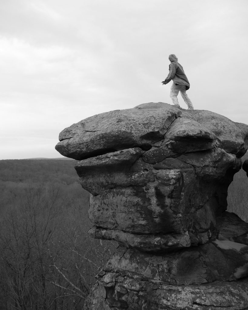 You stand below a rock balanced precariously on a cliff. A woman stands at the edge of the rock, back facing you, her jacket flaps in the sharp wind. A forest of bony trees stands below you stretching out to the horizon.